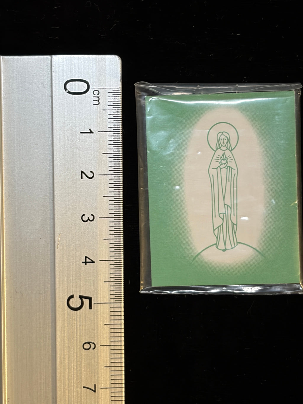 Green Scapular next to a ruler showing its size, about 3.8 x 4.9 cm