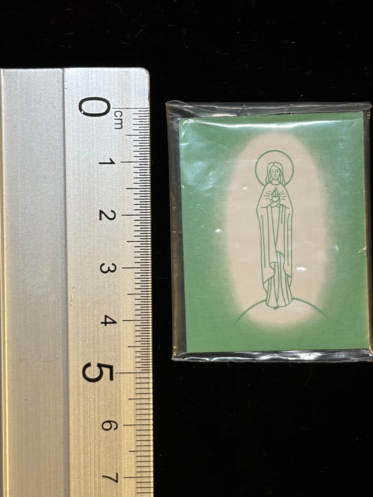 Green Scapular next to a ruler showing its size, about 3.8 x 4.9 cm
